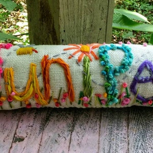 May include: A white pillow with the word "Juniper" embroidered in colorful thread. The letters are surrounded by flowers and leaves in shades of pink, yellow, blue, and green.