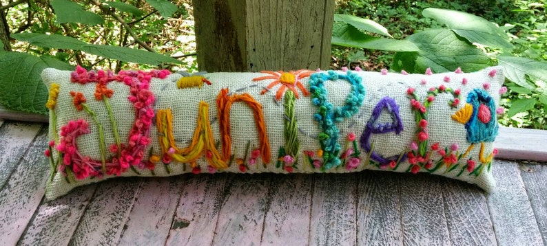 May include: A white pillow with the word "Juniper" embroidered in colorful thread. The letters are surrounded by flowers and leaves in shades of pink, yellow, blue, and green.