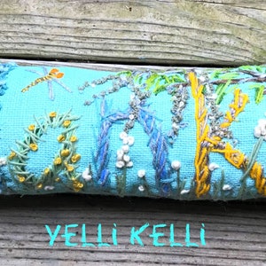 May include: Embroidered rectangular pillow with a light blue background. The pillow features a nature scene with a dragonfly, flowers, and a duck. The word "HOME" is embroidered on the pillow. The text "YELLI KELLI" is at the bottom.