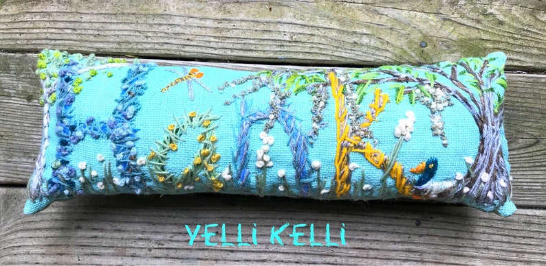 May include: Embroidered rectangular pillow with a light blue background. The pillow features a nature scene with a dragonfly, flowers, and a duck. The word "HOME" is embroidered on the pillow. The text "YELLI KELLI" is at the bottom.