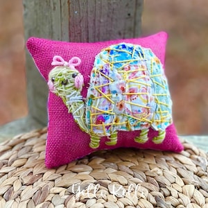 May include: A decorative pink pillow featuring a handcrafted turtle design. The turtle has a colorful shell made of fabric scraps and yellow stitching. The turtle's head and legs are green, and it has a pink bow. The pillow is on a woven surface.