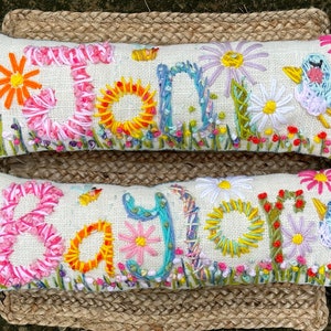 May include: Two embroidered pillow covers with the names "Jane" and "Sayler" spelled out in colorful thread. The pillows are decorated with flowers and birds.