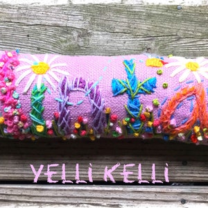 May include: A lavender-colored, rectangular decorative pillow with embroidered floral and cross designs. The pillow features a variety of colorful flowers, including daisies and other blooms, with the text "YELLI KELLI" at the bottom.