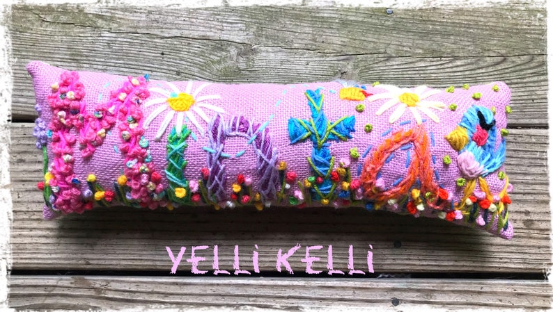 May include: A lavender-colored, rectangular decorative pillow with embroidered floral and cross designs. The pillow features a variety of colorful flowers, including daisies and other blooms, with the text "YELLI KELLI" at the bottom.