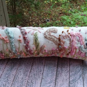 May include: A white linen pillow with embroidered floral designs in shades of pink, blue, and green. The pillow is decorated with a variety of flowers, including daisies, roses, and leaves.