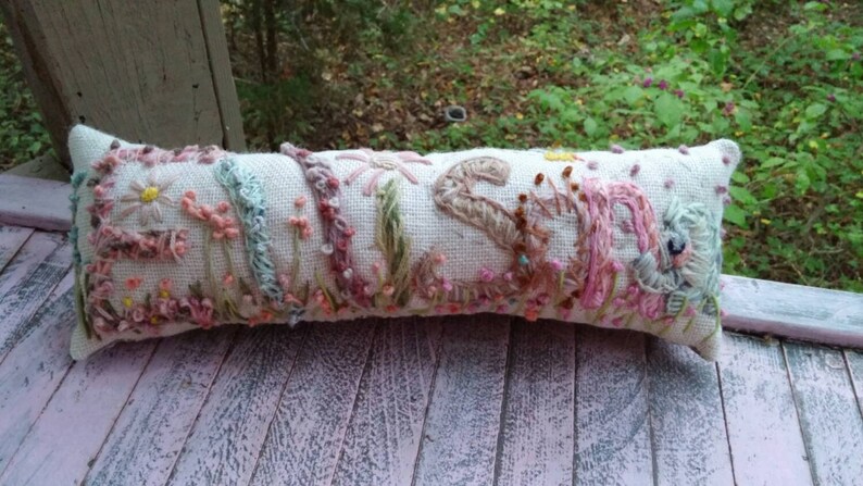 May include: A white linen pillow with embroidered floral designs in shades of pink, blue, and green. The pillow is decorated with a variety of flowers, including daisies, roses, and leaves.