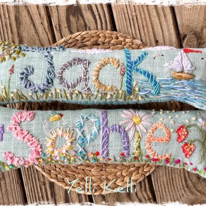 May include: Two embroidered rectangular pillows with names, "Jack" and "Sophie", on a light blue background. The Jack pillow features a sailboat and tree. The Sophie pillow has flowers and a bee. Both are on a woven mat.