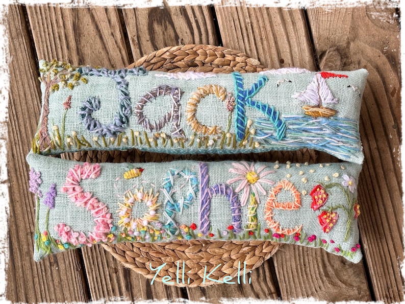 May include: Two embroidered rectangular pillows with names, "Jack" and "Sophie", on a light blue background. The Jack pillow features a sailboat and tree. The Sophie pillow has flowers and a bee. Both are on a woven mat.