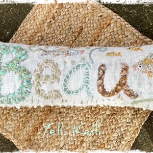 May include: Embroidered rectangular pillow with the word "BEACH" in textured, colorful stitching. The pillow features fish and aquatic plant designs. It rests on a woven, natural-toned mat.