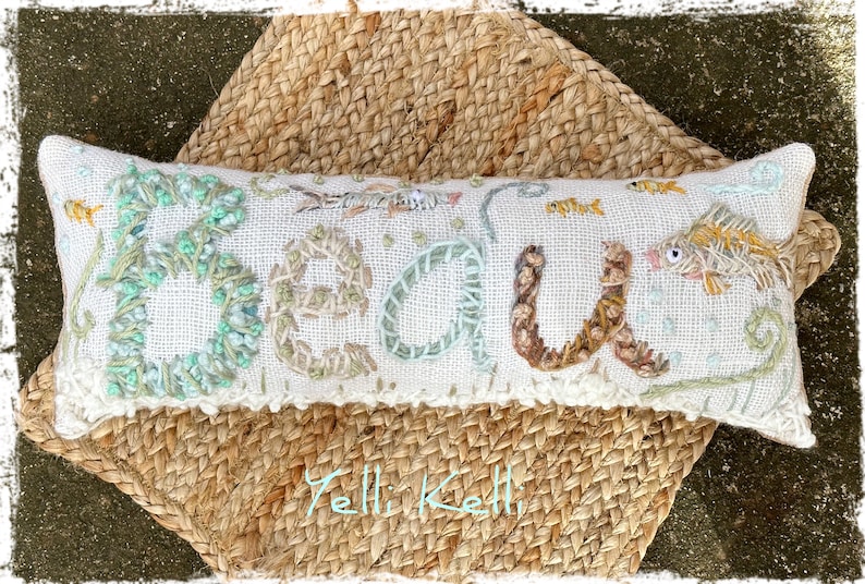 May include: Embroidered rectangular pillow with the word "BEACH" in textured, colorful stitching. The pillow features fish and aquatic plant designs. It rests on a woven, natural-toned mat.