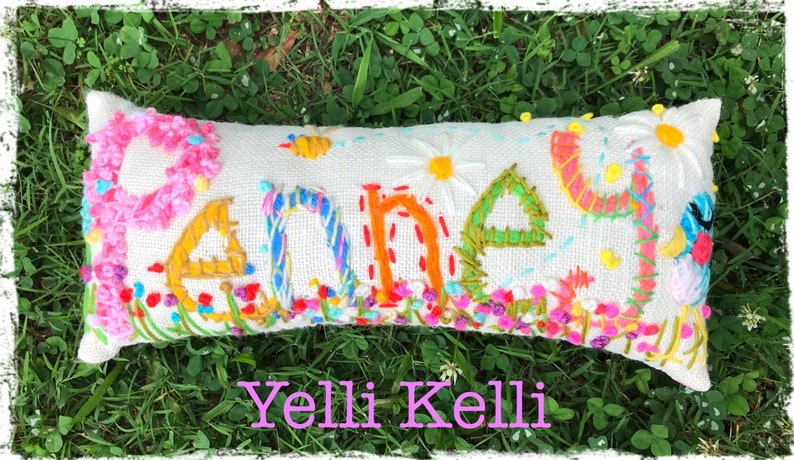 May include: A white pillow with the word "Poney" embroidered in colorful thread. The letters are surrounded by embroidered flowers and a bee. The pillow is lying on green grass. The words "Yelli Kelli" are written in pink below the pillow.