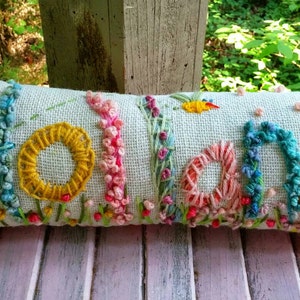 May include: A long, rectangular pillow with the word "Holland" embroidered in colorful thread. The letters are surrounded by embroidered flowers and leaves.