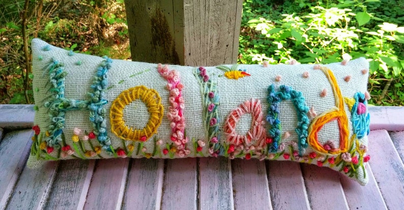 May include: A long, rectangular pillow with the word "Holland" embroidered in colorful thread. The letters are surrounded by embroidered flowers and leaves.