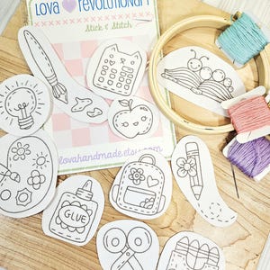May include: A selection of stick and stitch embroidery patches featuring school-related motifs such as a paintbrush, apple, and backpack. The image also includes embroidery threads, a wooden hoop, and the text "Lova Revolutionary Stick & Stitch."
