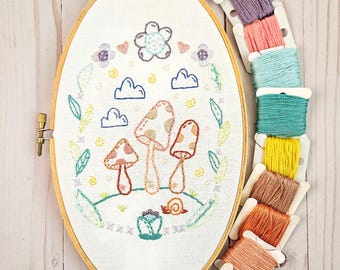 May include: A white embroidery hoop featuring a colourful embroidered design of mushrooms, clouds and a snail. The hoop is surrounded by spools of embroidery floss in various colours.