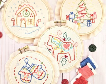 May include: Four embroidered Christmas ornaments in wooden hoops. The ornaments feature a gingerbread house, a Christmas tree, a Dala horse, and a pair of mittens. The ornaments are on a white wooden surface with red and green buttons and spools of embroidery floss.