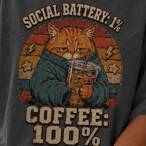 Könnte beinhalten: Ein anthrazitfarbenes T-Shirt mit einer Grafik einer orangefarbenen Katze in einem blauen Pullover, die einen Eiskaffee hält. Der Text lautet SOCIAL BATTERY: 1% COFFEE: 100% vor einem kreisförmigen Sonnenuntergangshintergrund mit Sternen und Blitzen.