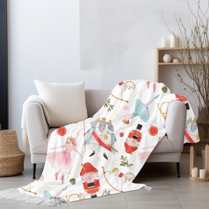 May include: White throw blanket with a repeating pattern of watercolor illustrations. The design includes ballerinas, nutcrackers, and other festive elements in shades of red, pink, blue, and gold. The blanket is draped over a beige sofa.