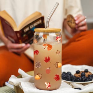 May include: A clear glass tumbler with a bamboo lid and a glass straw. The tumbler is decorated with autumn-themed stickers, including a fox, leaves, and acorns. The tumbler is filled with a light-colored beverage.