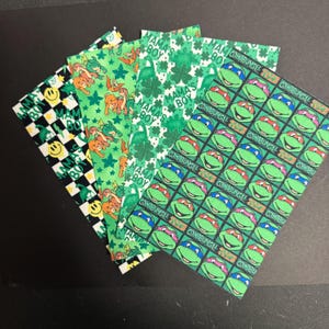 May include: A collection of patterned fabric squares. Designs include a black and white checkerboard with smiley faces, green dinosaurs, shamrocks, and Teenage Mutant Ninja Turtles. The text "MAMA'S BOY" and "COWABUNGA" are visible.