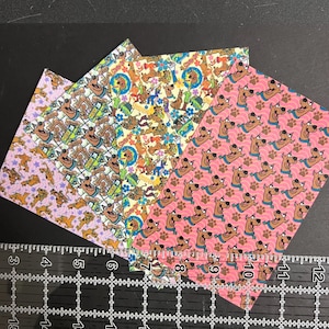May include: Three sheets of decorative paper featuring a cartoon dog with the text "Scooby Dooby Doo" printed on them. The paper is pink, yellow, and purple with a variety of patterns.