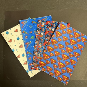 May include: Four square fabric pieces with Superman-themed patterns. Designs include the Superman logo, Superman in flight, and a pattern of stars and Superman logos. Colors are blue, red, yellow, and white.