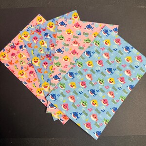 May include: A collection of colorful patterned papers featuring cartoon sharks in various colors, including yellow, pink, blue, and green. The papers also have musical notes and the word "Doo" printed on them. The papers are arranged in a fan shape.