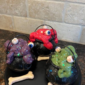 May include: Three Halloween-themed cauldrons with black bases and handles, each filled with colorful, textured goo and a large eyeball. The cauldrons are decorated with glitter and sit on a black tray with bone-shaped accents.