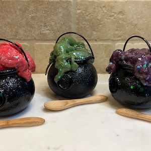 May include: Three small black cauldrons with different colored slime inside. Each cauldron has a handle and is sitting on a white surface. There are three wooden spoons next to the cauldrons.