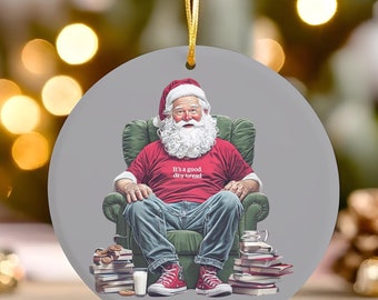 May include: A round Christmas ornament featuring a cartoon Santa Claus wearing a red shirt with the text "It's a good day to be a rebel" sitting in a green armchair with a stack of books and a mug.