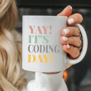 May include: White ceramic mug with the text "YAY! IT'S CODING DAY!" in colorful letters. The mug is being held by a person.