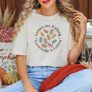 May include: A white t-shirt with a graphic of colorful leaves in a circle. The text "Leaves are Falling Knowledge is Calling" is written around the leaves.