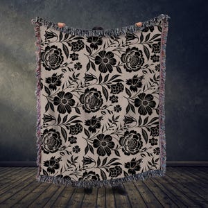 May include: A beige throw blanket with a black floral pattern. The design features large flowers, leaves, and smaller blossoms. The blanket has a fringed edge and is held up against a dark background.