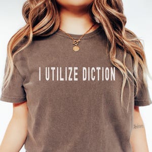 May include: A brown t-shirt with the phrase "I Utilize Diction" printed in white, centered on the front. The shirt has short sleeves and a crew neck. The model is wearing a gold necklace.