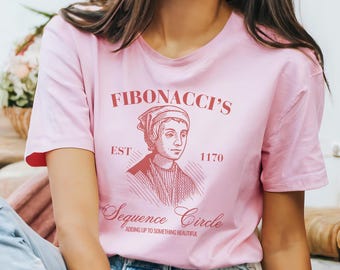 May include: A light pink t-shirt featuring a graphic of a man with the text "FIBONACCI'S" above the image, and "Sequence Circle" below. The shirt also includes the text "EST 1170" and "Adding up to something beautiful".