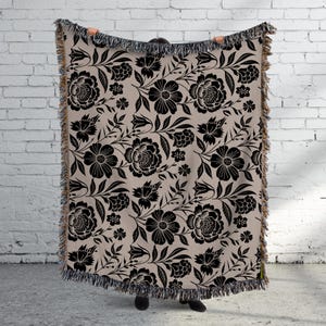 May include: A beige throw blanket with a black floral pattern. The design features large flowers, leaves, and smaller floral accents. The blanket has a fringed edge.