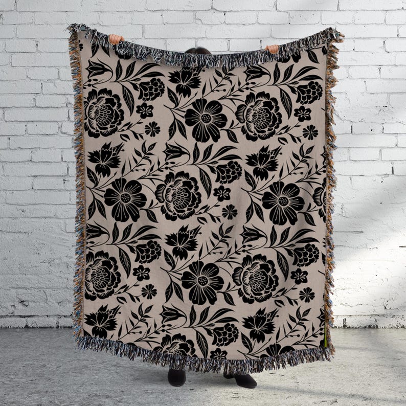 May include: A beige throw blanket with a black floral pattern. The design features large flowers, leaves, and smaller floral accents. The blanket has a fringed edge.