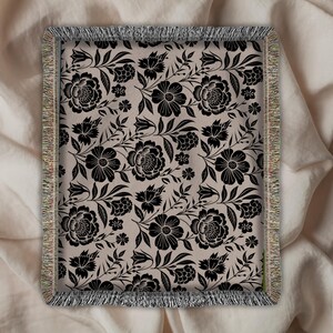 May include: A rectangular throw blanket with a floral pattern. The design features black flowers and leaves on a beige background. The blanket has a fringed border in shades of beige, tan, and green. The floral pattern is a repeating design.