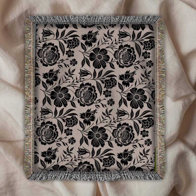 May include: A rectangular throw blanket with a floral pattern. The design features black flowers and leaves on a beige background. The blanket has a fringed border in shades of beige, tan, and green. The floral pattern is a repeating design.