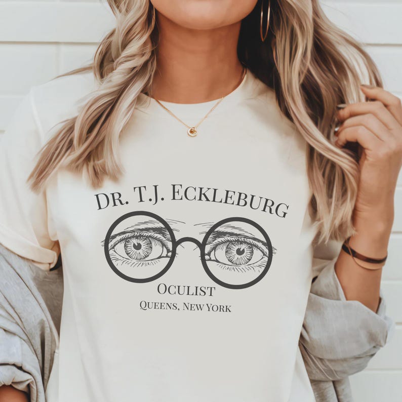 以下が含まれることがあります： クリーム色のTシャツ。上部に「DR. T.J. ECKLEBURG」の文字、丸いメガネフレームの中に2つの目のグラフィック。グラフィックの下には「OCULIST」と「QUEENS, NEW YORK」の文字。
