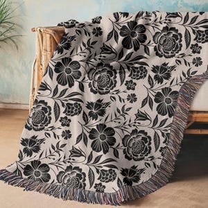 May include: A beige throw blanket with a black floral pattern, draped over a light-colored sofa. The blanket features a repeating design of large flowers, leaves, and smaller floral accents. The edge of the blanket has a fringed trim.