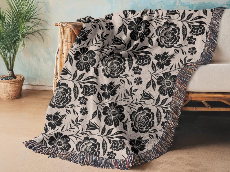May include: A beige throw blanket with a black floral pattern, draped over a light-colored sofa. The blanket features a repeating design of large flowers, leaves, and smaller floral accents. The edge of the blanket has a fringed trim.