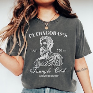 May include: Dark gray t-shirt with a white graphic of Pythagoras and the text "Pythagoras's Triangle Club" with the phrase "Where Every Side Has a Story". The shirt has a vintage, worn look.