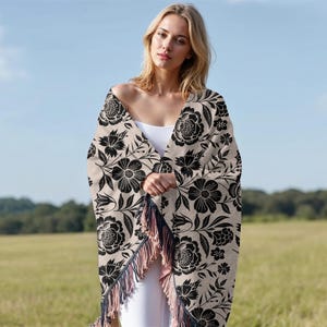 May include: A beige shawl with a black floral pattern and fringe. The shawl is draped over the shoulders. The background is a field and a blue sky.