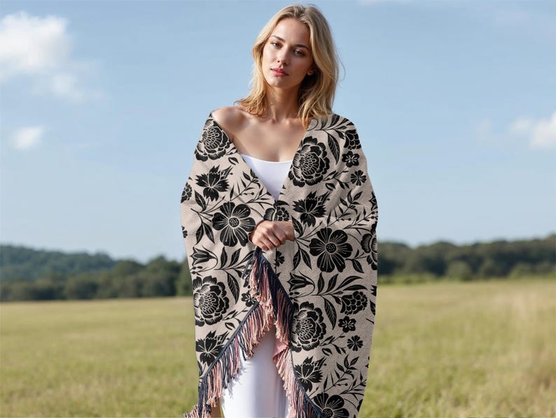 May include: A beige shawl with a black floral pattern and fringe. The shawl is draped over the shoulders. The background is a field and a blue sky.