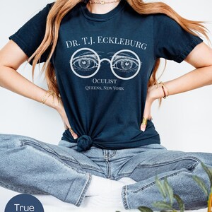 以下が含まれることがあります： 白文字で「DR. T.J. ECKLEBURG OCULIST QUEENS, NEW YORK」と書かれたネイビーブルーのTシャツ。デザインは、詳細な目のイラストが描かれたメガネが特徴です。シャツはウエストで結ばれています。