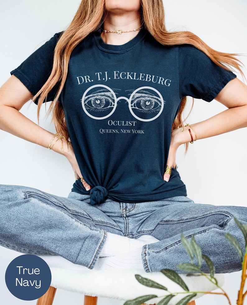 以下が含まれることがあります： 白文字で「DR. T.J. ECKLEBURG OCULIST QUEENS, NEW YORK」と書かれたネイビーブルーのTシャツ。デザインは、詳細な目のイラストが描かれたメガネが特徴です。シャツはウエストで結ばれています。