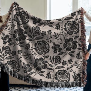 May include: A beige throw blanket with a black floral pattern, including large peony-like flowers and smaller blossoms. The blanket has a fringed edge and is held up by two people.