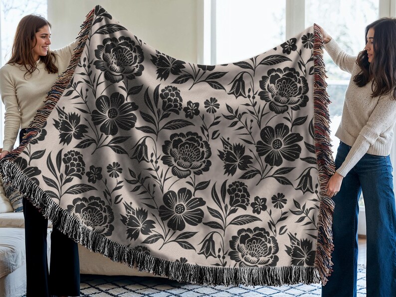 May include: A beige throw blanket with a black floral pattern, including large peony-like flowers and smaller blossoms. The blanket has a fringed edge and is held up by two people.