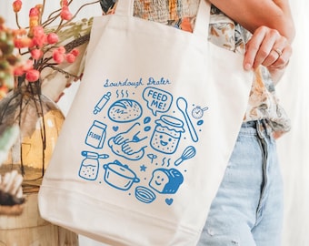 May include: Cream-colored tote bag with blue illustrations of baking tools and food items. The words "Sourdough Dealer" and "FEED ME!" are printed on the bag. The bag has a rectangular shape and two handles.